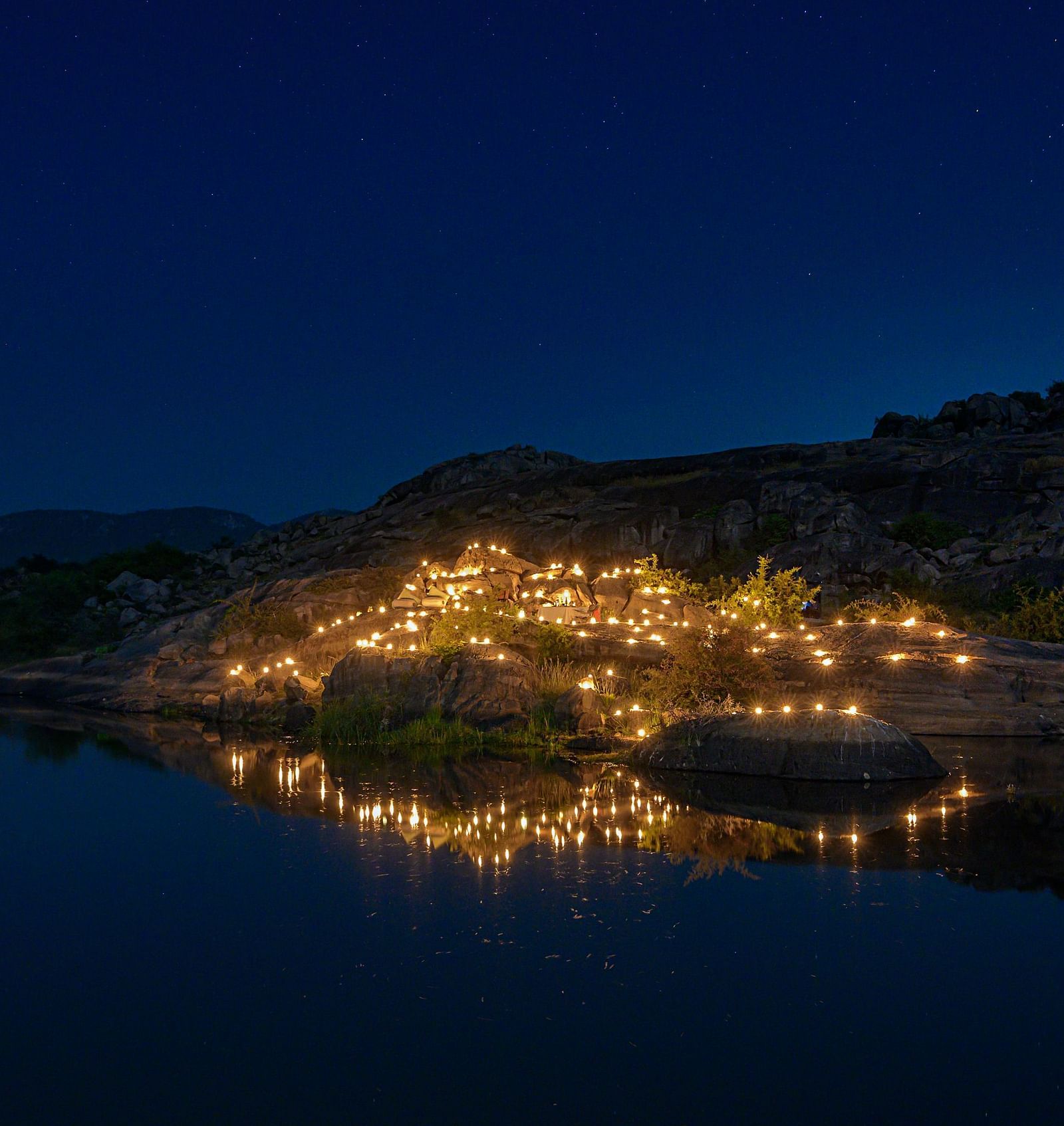 An image of an outdoor dining arrangement near a lake over viewing the mountains with bright lighting and greenery around - Utsav Camp Sariska