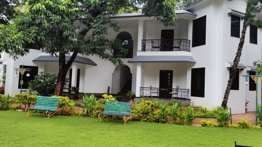 An exterior view of a white multi-story building featuring balconies, surrounded by trees and a green grass lawn at Amantra Shilpi Resort, Udaipur.