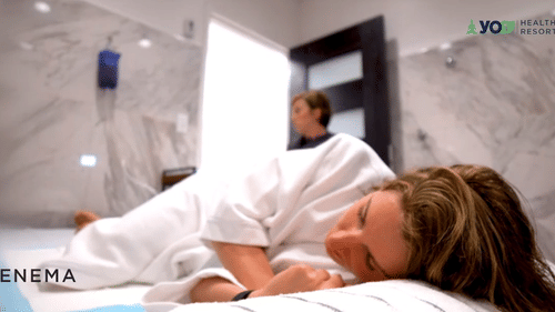 A person in a white robe lying on their side on a bed, with another person in the background at YO1 Longevity & Health Resorts