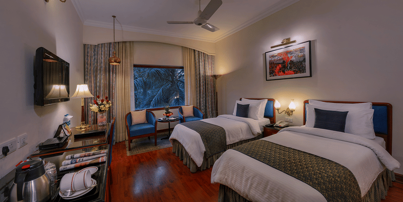 A Premiere Room at The Clarks Varanasi featuring twin beds, a ceiling fan, a wall-mounted television, and a seating area at The Clarks Varanasi.