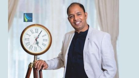 A man in a white jacket standing in front of a wall clock, pointing at it with a confident expression.