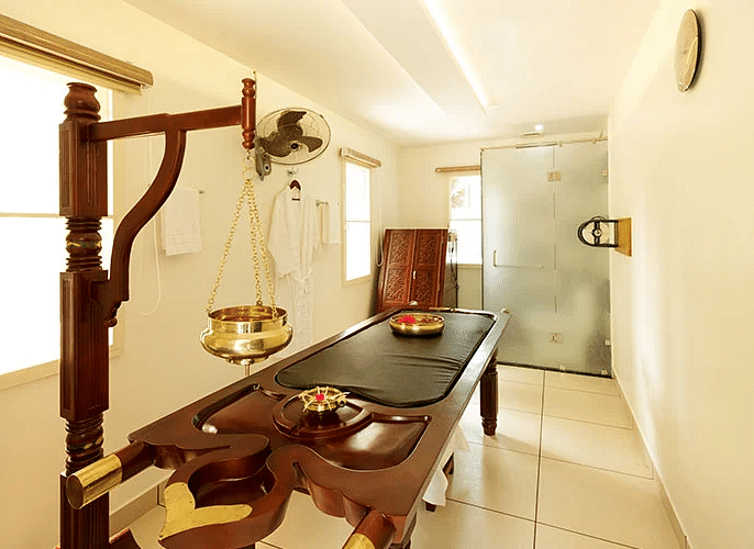 A traditional wooden massage table in a white room with natural light, suggesting a treatment area at Lhasa Ayurveda & Wellness Resort.