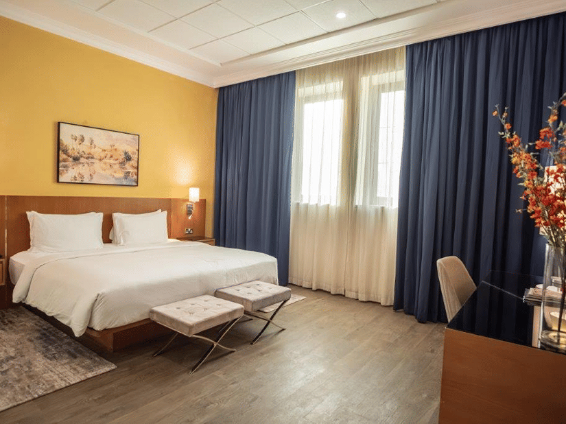 Well-lit bedroom with a king-size bed, desk, chair, and decorative elements at La Maison Hotel, Doha.