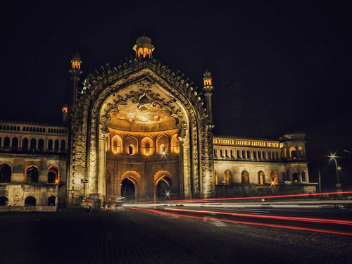 A historic, grand arched gateway of the Rumi Darwaza in Lucknow is illuminated at night, with light trails from cars on the street in front of it.