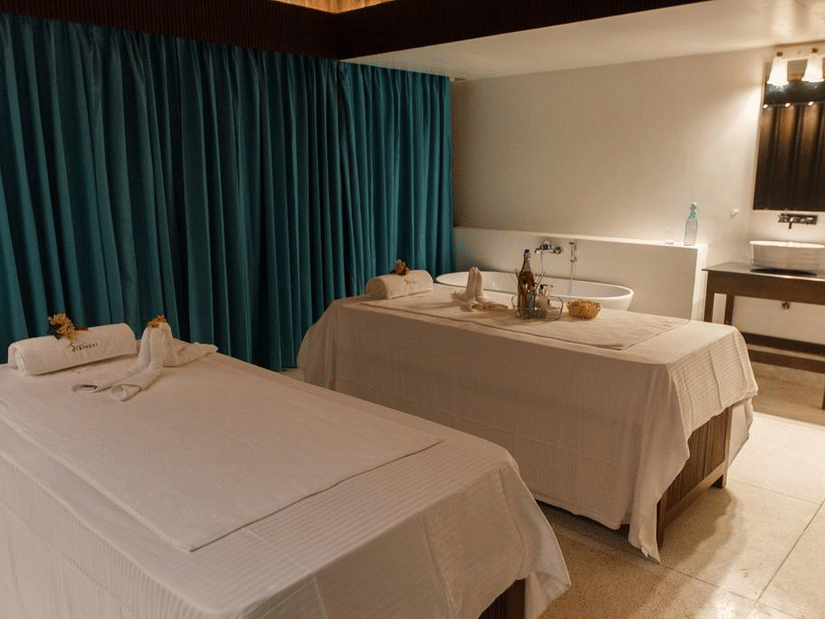 Interior of Svarasyaa Wellness, a Port Blair spa at Symphony Samudra Beachside Jungle Resort And Spa featuring 2 neatly made massage tables with white linens, folded towels, and a bathtub at the side of the room.