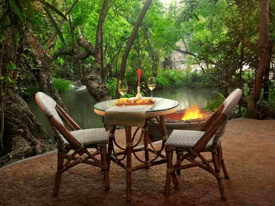 Outdoor dining table with wicker chairs set beneath a canopy of trees at Amanvana Spa Resort, Coorg.