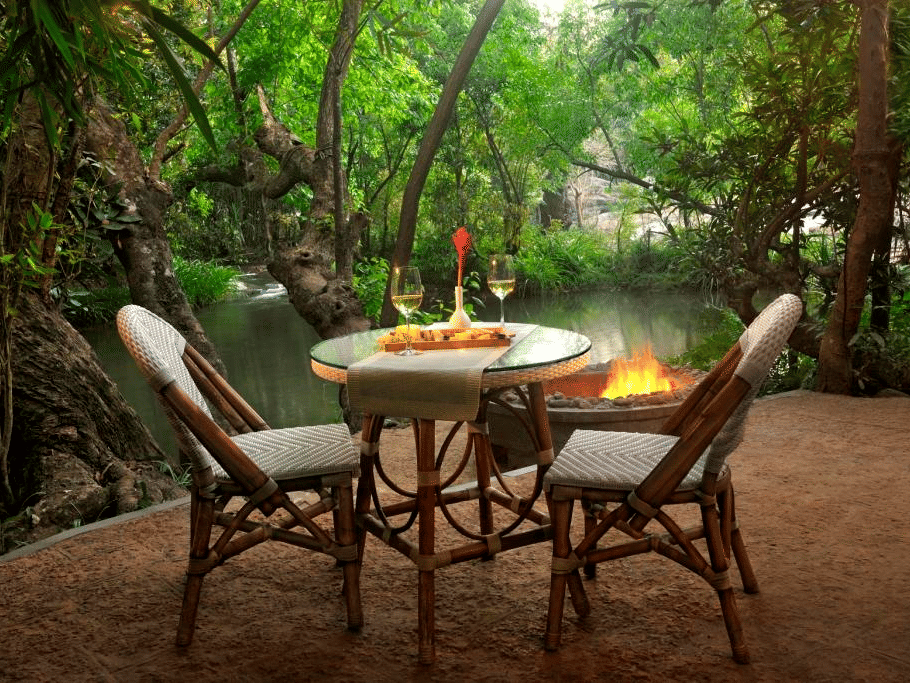 An outdoor dining setup with chairs for a couple, a small table, and a bonfire setup near a tranquil river in the forest at the Amanvana Spa Resort, Coorg - Luxury Resort