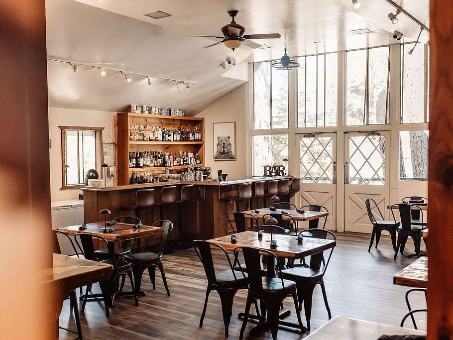 A cafe at Shaver Lake Village Hotel with wooden tables, metal chairs, large windows, and warm lighting, creating an inviting ambiance.