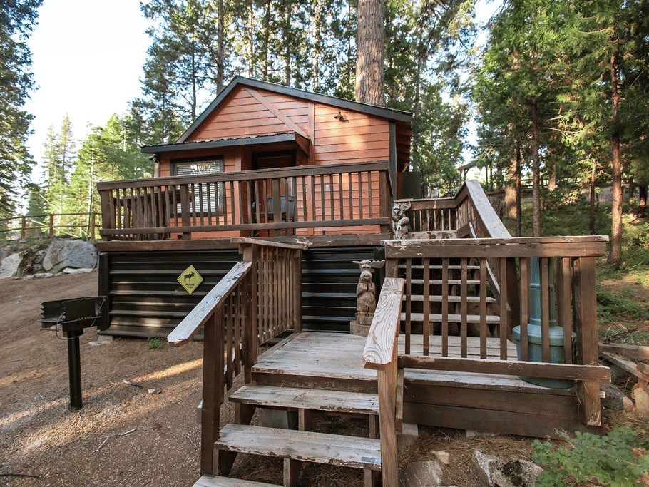 A Cabin at Shaver Lake Village Hotel with a wooden deck, stairs, and a grill, surrounded by tall pine trees.