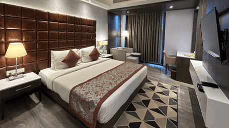 A view of a hotel room showing a bed with a dark headboard, a bedside lamp, and a television on the wall | Sand Heaven