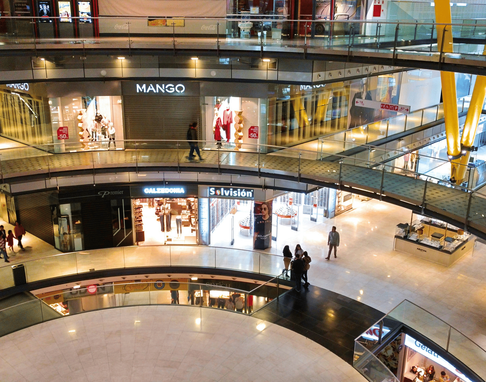 Interior view of a modern, multi-level shopping centre with glass railings, bright lighting, and various shops visible.