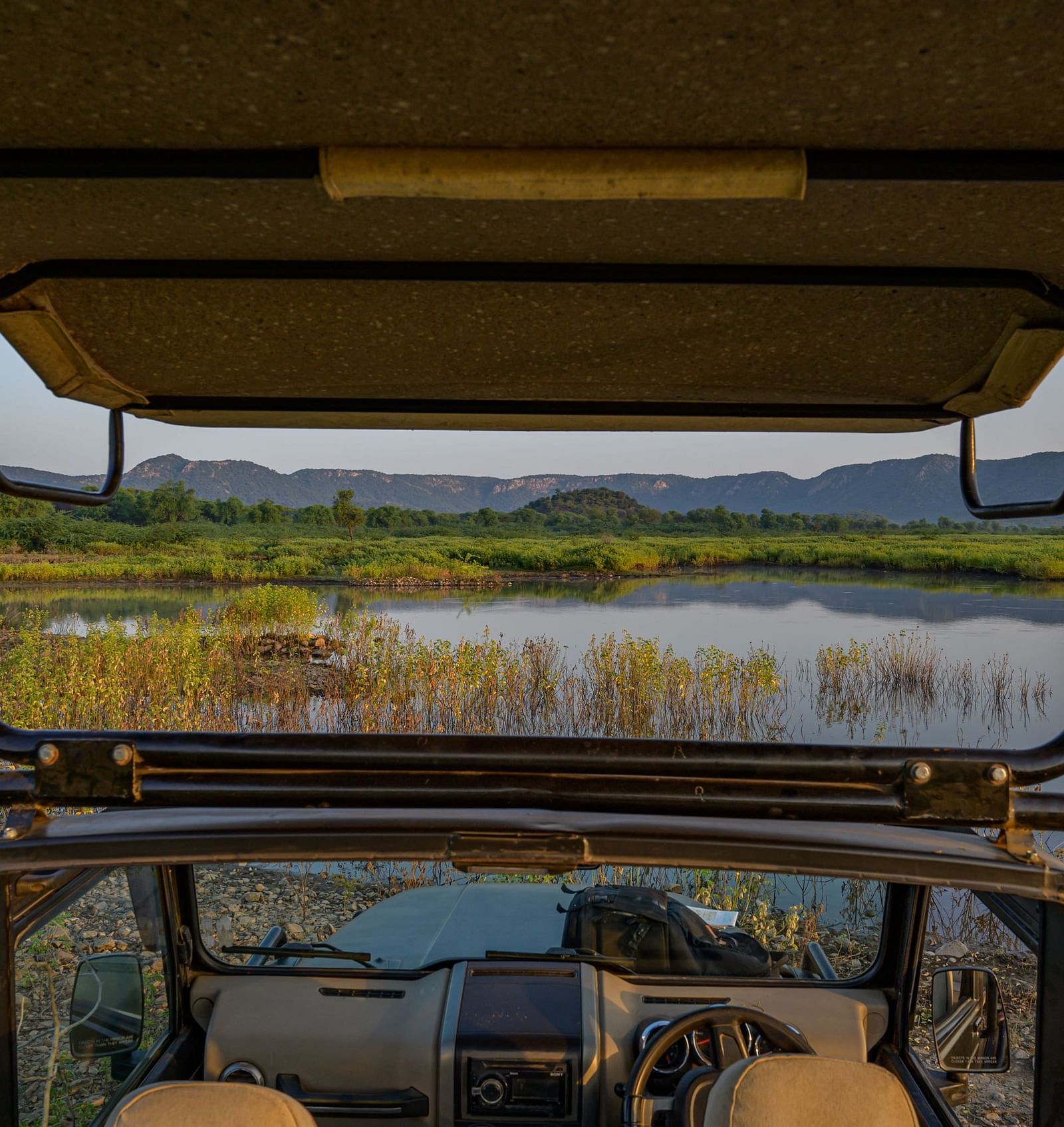 An image of the inside of a jeep over viewing a lake and the mountains -0 An image of a jeep parked amidst the rocky mountains - Utsav Camp Sariska