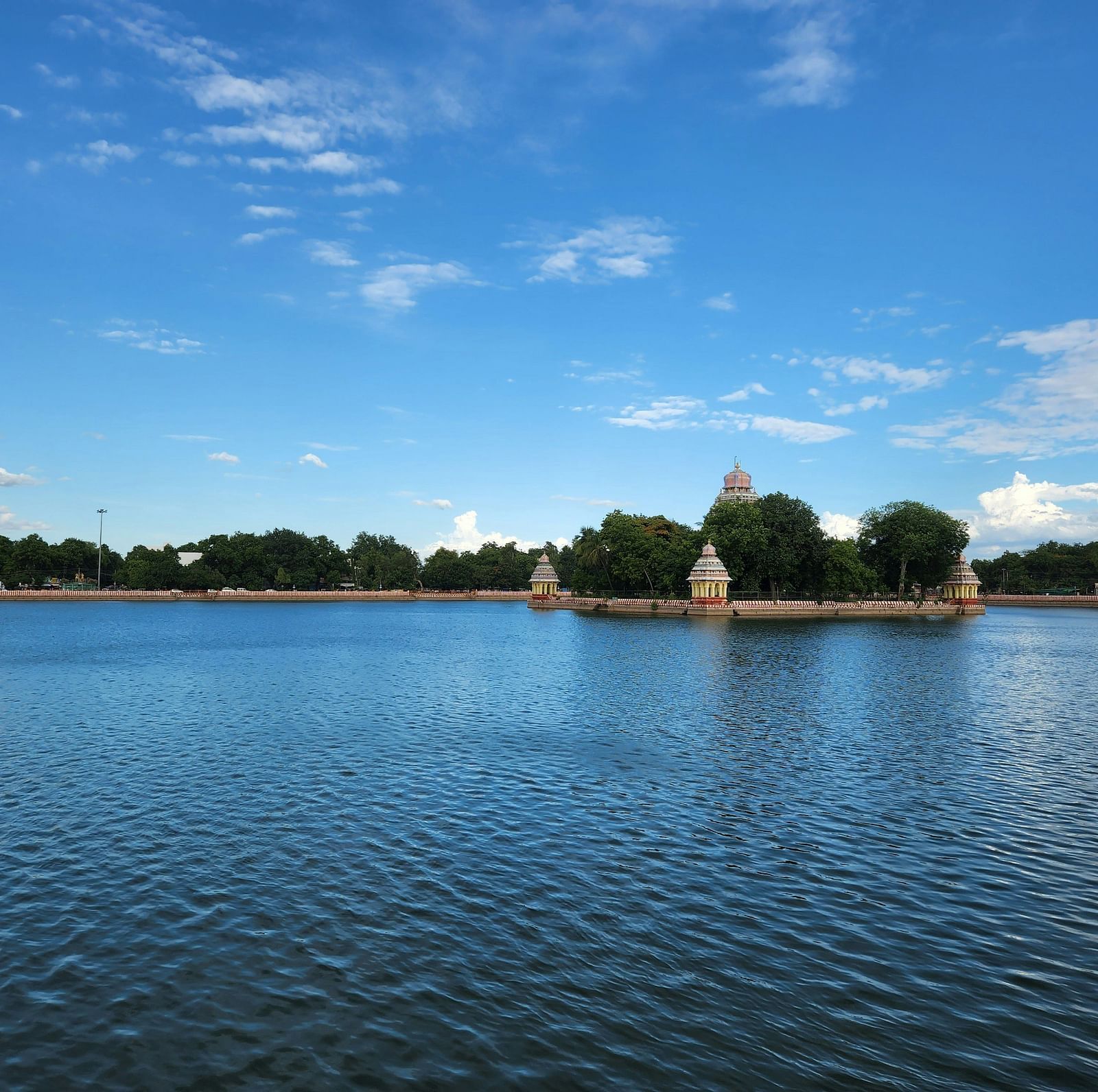 A scenic view of a temple tank, surrounded by trees with a blue sky and calm waters.
