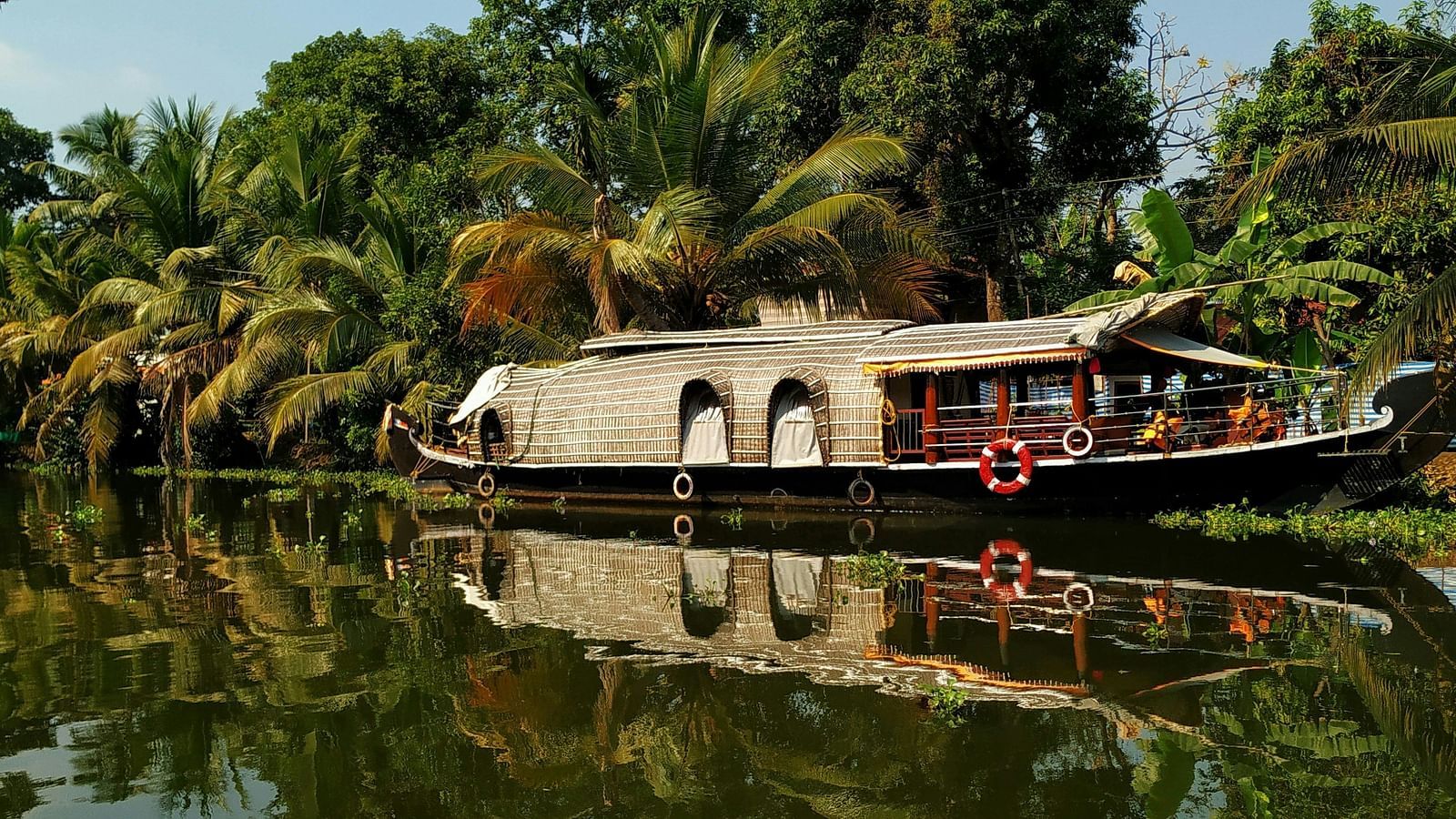 A traditional houseboat with a thatched roof is floating on the calm backwaters, flanked by lush greenery and palm trees reflecting in the water.