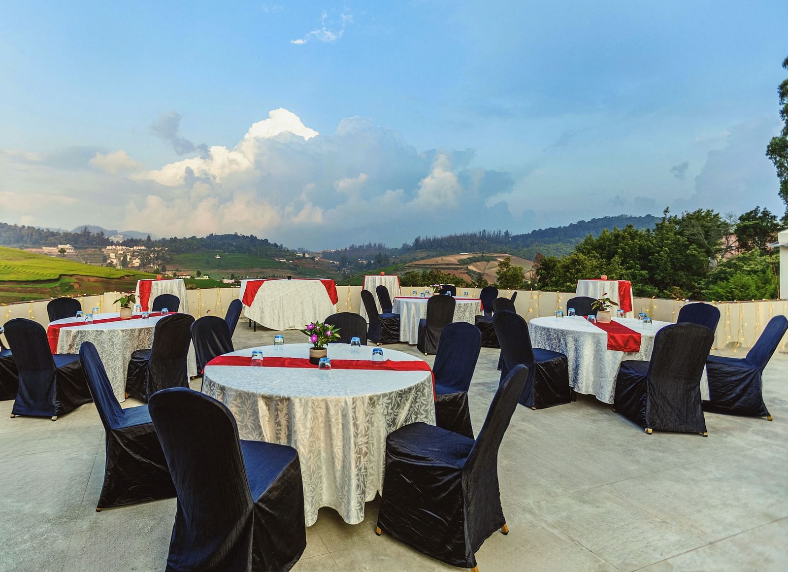 Rooftop event setup at Mango Hill Shola, Ooty, featuring round tables covered in white linens with red and black accents, surrounded by black banquet chairs, overlooking lush green hills and a dramatic cloudy sky.