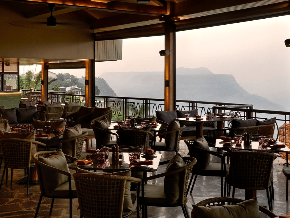 Restaurant dining area with rattan furniture and ambient lighting, featuring large windows with a stunning panoramic view of a mountain valley.