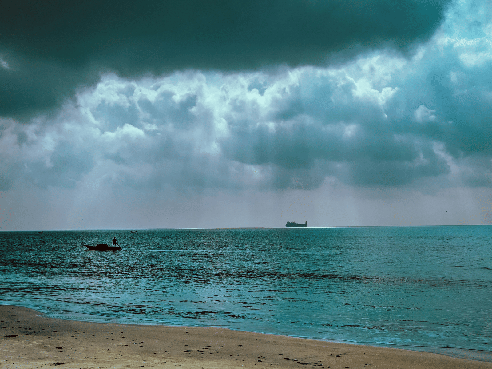 A small boat with a person near a sandy beach faces a dark, cloudy sky with sun rays breaking through, while a large cargo ship sits on the horizon of the teal ocean.