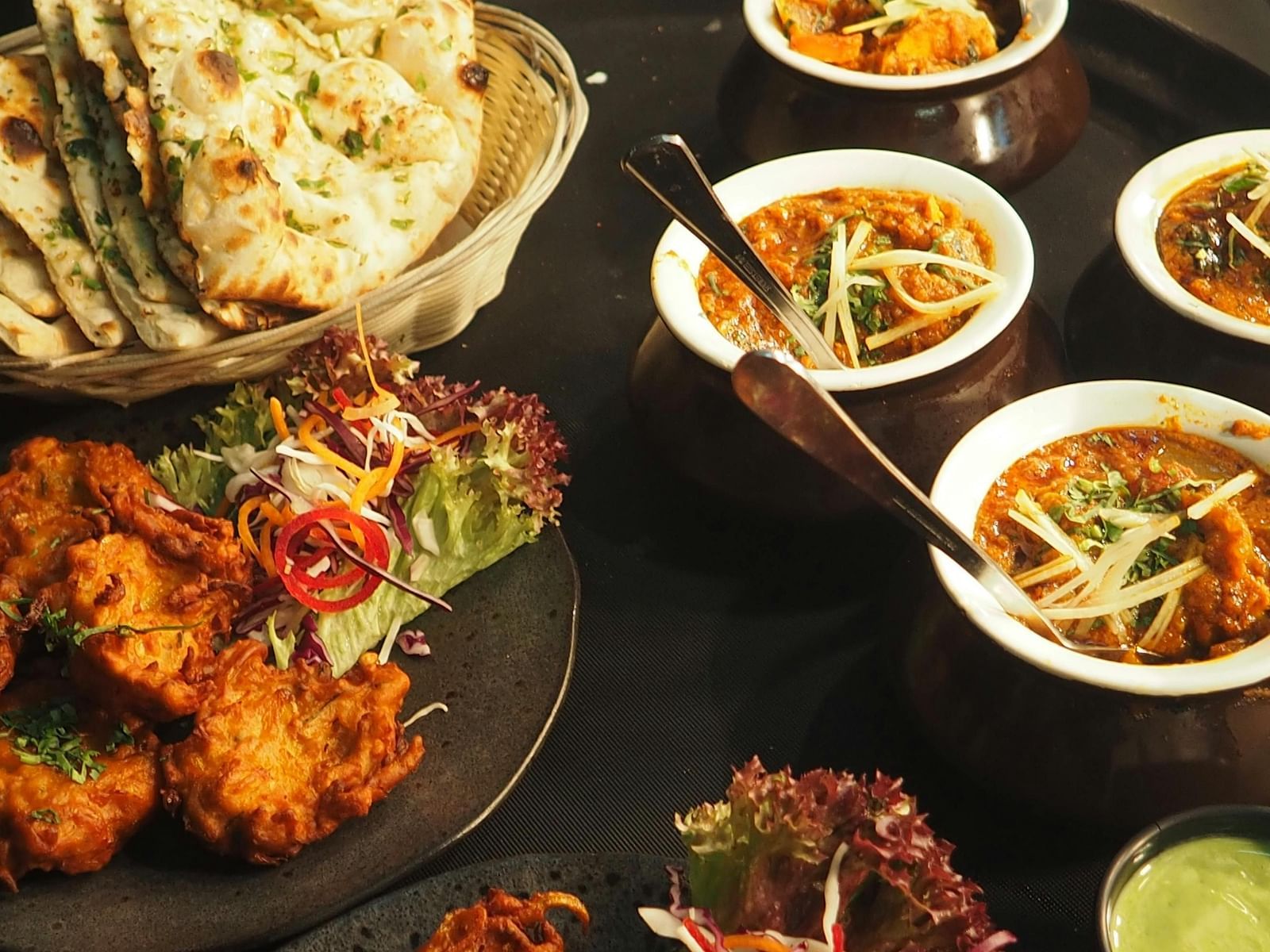 A spread of Indian dishes, including naan, pakora, and curries, is presented on a dark surface.