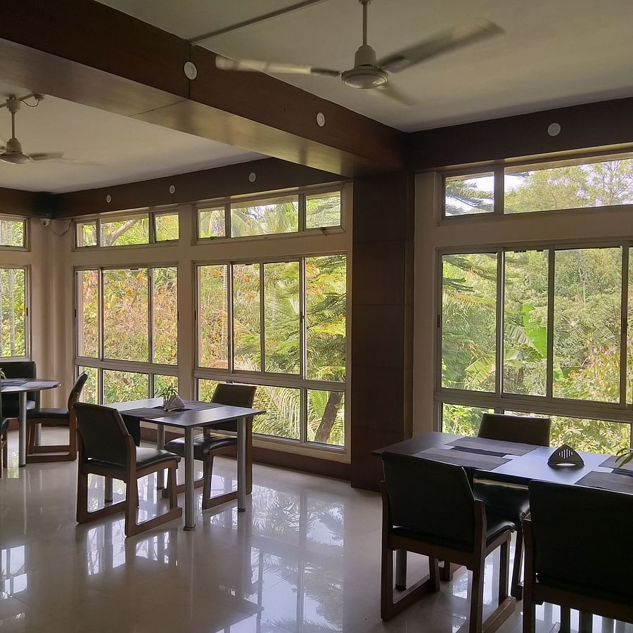 alt-text A bright and airy hotel dining space with tables set for service, surrounded by large windows offering views of greenery.