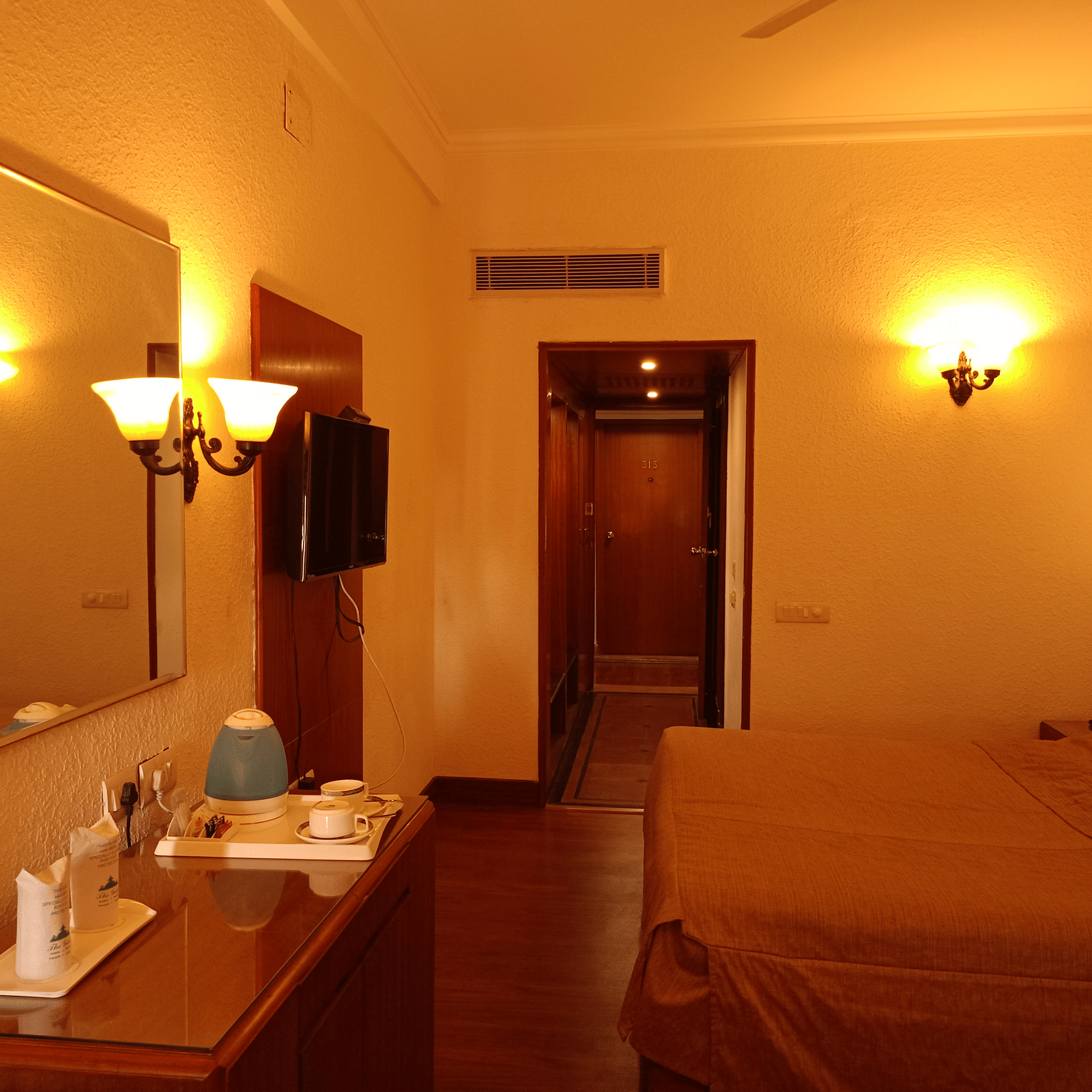 Room interior at Hotel Gold, Panipat with a bed, lamps, desk, and a mirror near the entrance.
