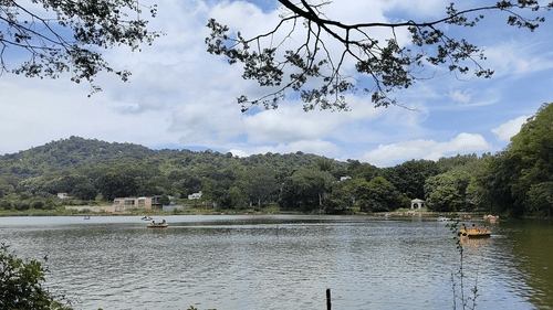 A calm lake surrounded by green hills and trees under a bright sky with scattered clouds. People are paddling small boats on the water, and a white pavilion sits near the lake’s edge.