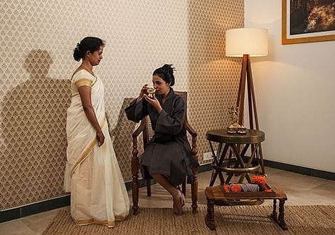 Two people in light-colored clothing standing in a room with a lamp and chairs - Shreyas Retreat