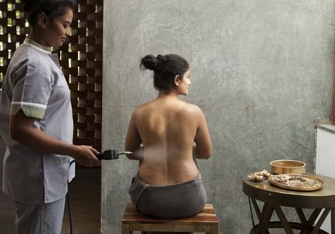 A person receiving a treatment on their back, with another person standing nearby, indoor setting - Shreyas Retreat