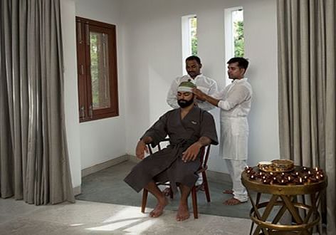 A person seated in a chair, wearing a dark garment with a white headdress, in a room with a window - Shreyas Retreat