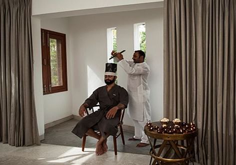 A person seated in a chair, wearing a dark garment with a white headdress, in a room with a window - Shreyas Retreat