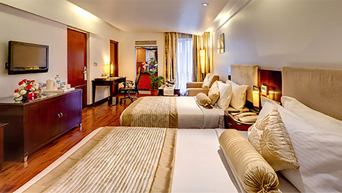 Side view of a Premium Room at Hotel Hindusthan International Select Bengaluru with twin beds, wall-mounted TV, wooden flooring, and warm ambient lighting.