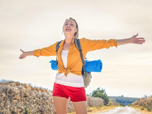 a woman smiling, with a backpack standing with her arms open outside on a sunny day