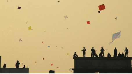 Yellow evening sky filled with numerous colourful kites flying above terracotta rooftops and distant cityscape at dusk during Jaipur festival