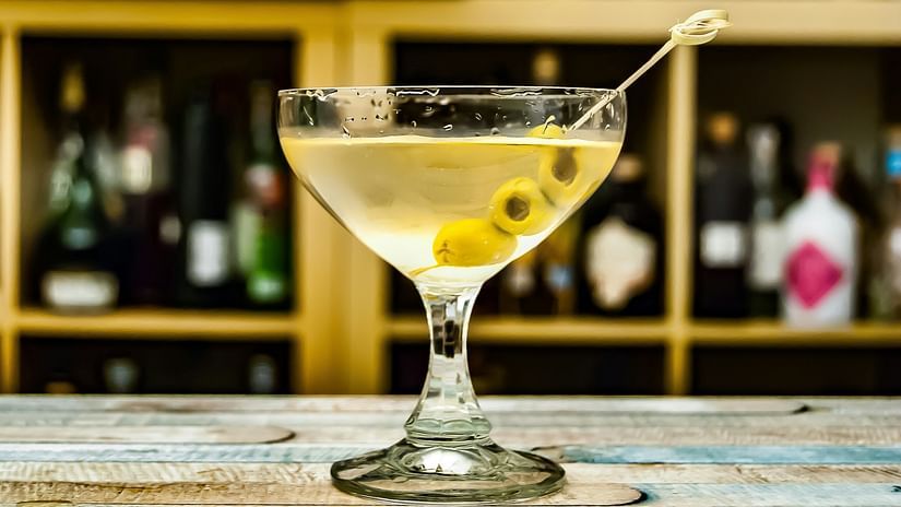 Cocktail glass on a bar counter, filled with a drink and garnished with olives, bottles in background.
