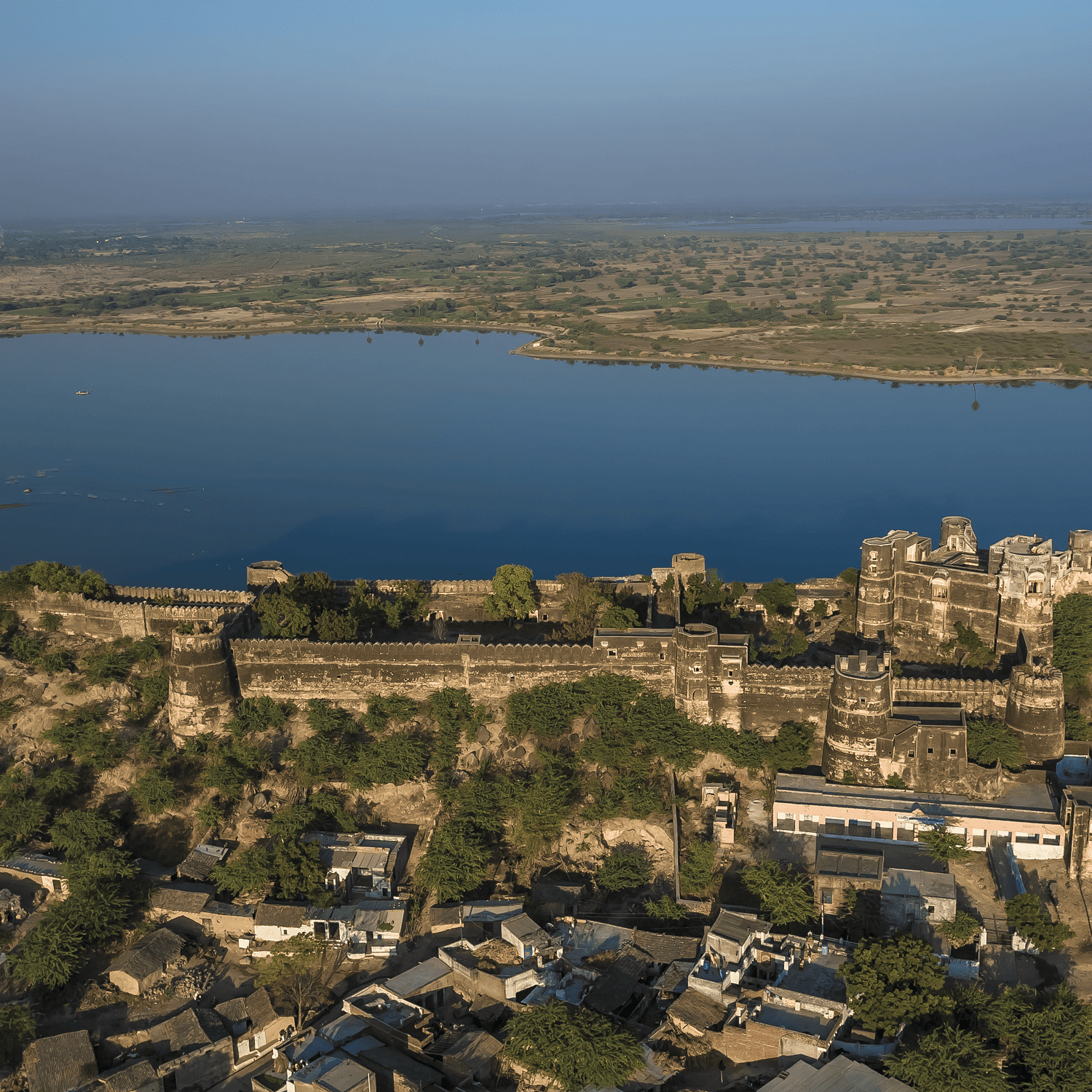 An aerial view of a sprawling fort complex on a hill, overlooking a large body of water and a hazy landscape stretching to the horizon- Shahpura Bagh.