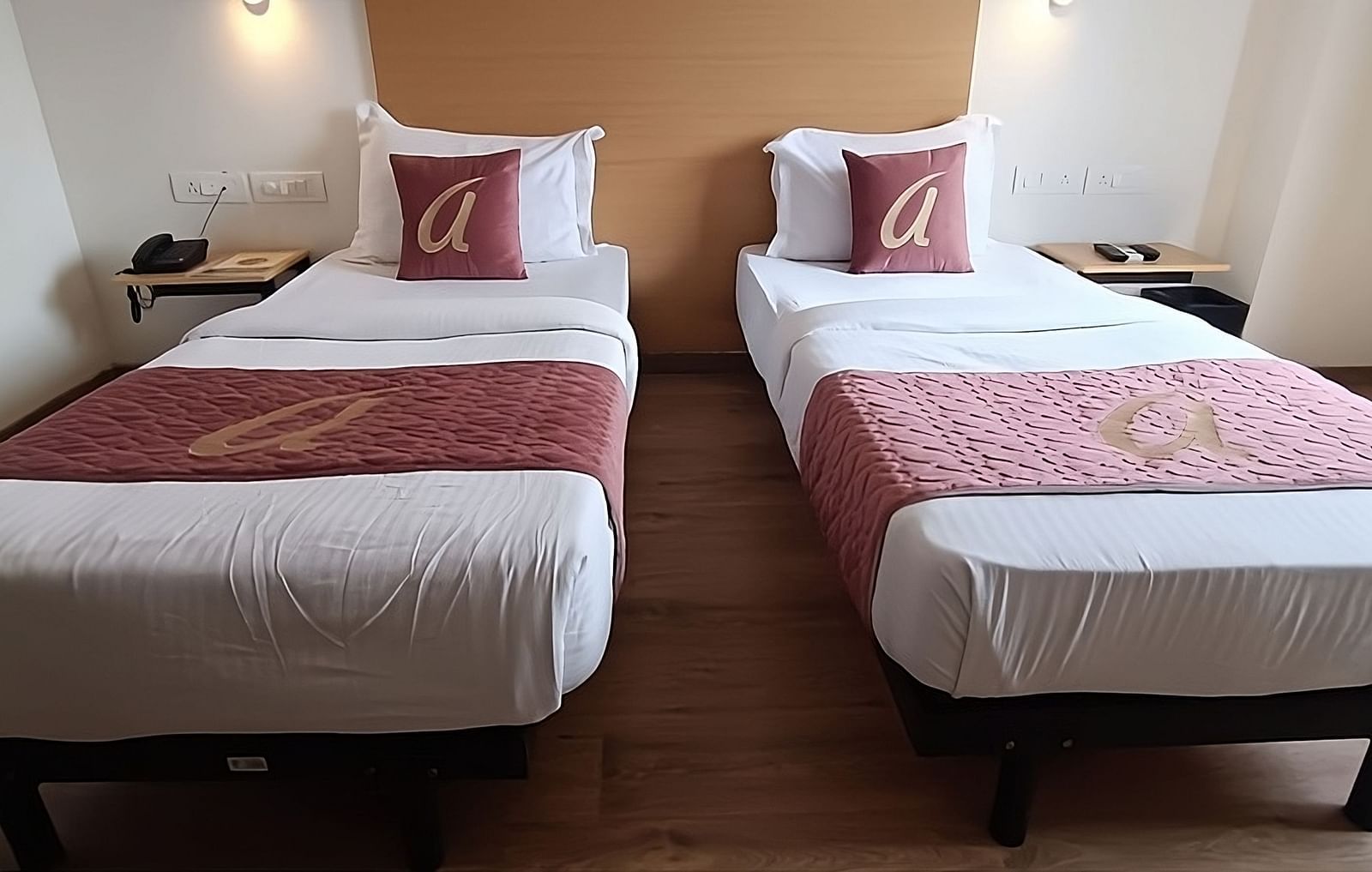 A well lit  bedroom with two single beds and red cushions at Astra Hotels and suits, Sarjapura.