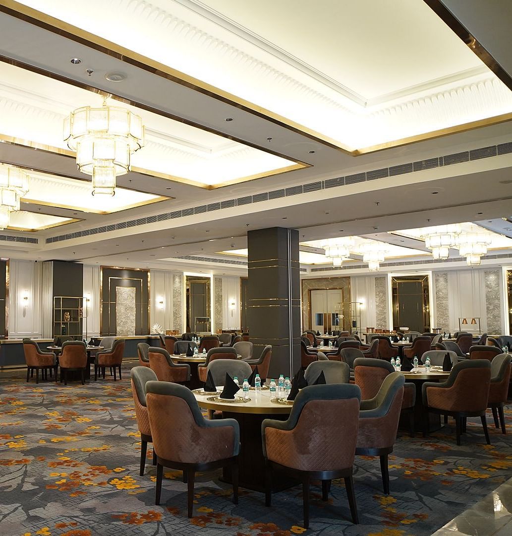 An elegant banquet hall at Nirvana Luxury Hotel, Ludhiana, featuring round tables with plush chairs on a patterned carpet, illuminated by multiple tiered chandeliers and recessed lighting.