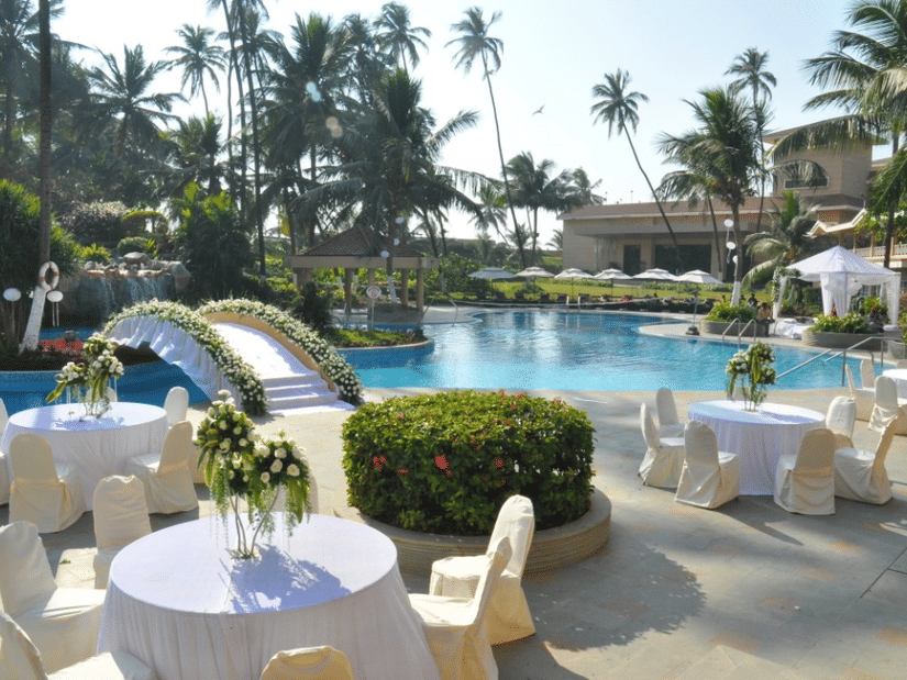 A poolside dining area at The Retreat Hotel and Convention Centre, with tables chairs, a bridge, with palm trees and a building in the background.