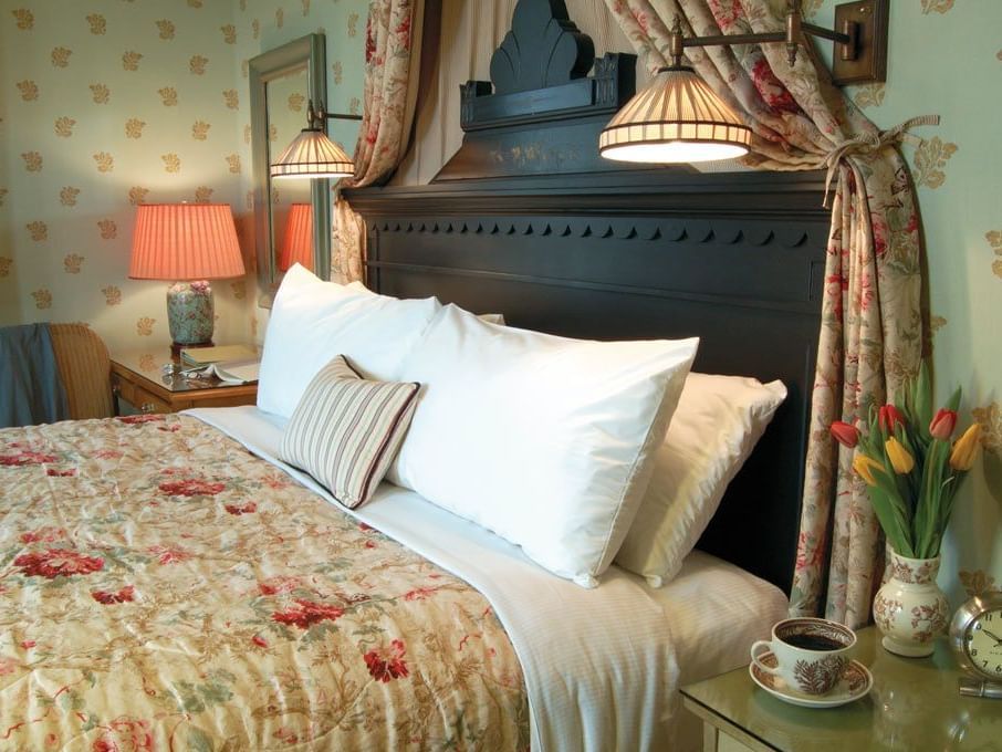 A bedroom at the Tallman Hotel contains a bed with a floral comforter and a dark wooden headboard.
