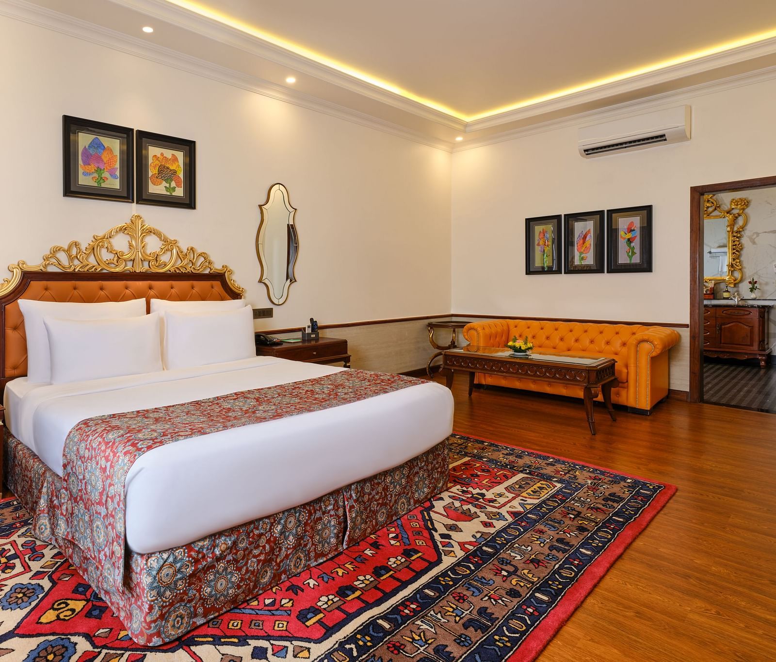 a luxurious suite, where a sofa, a table and a grand bed sits beneath a decorative headboard and artwork on the wall with two mirrors