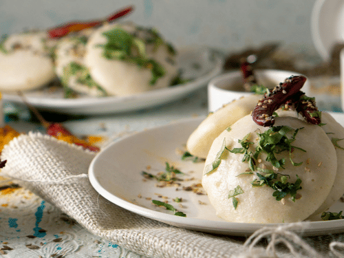 Plates of food served on a dining table with idlis, cutlery, bowls, and table setting visible.