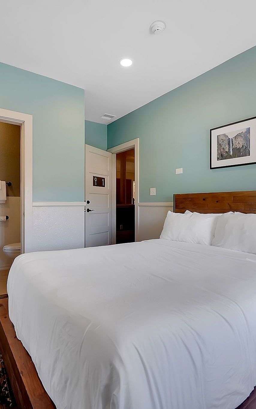 The Groveland Hotel's compact room features sage green walls, a wooden bed with crisp white bedding, and a pedestal sink with mirror.