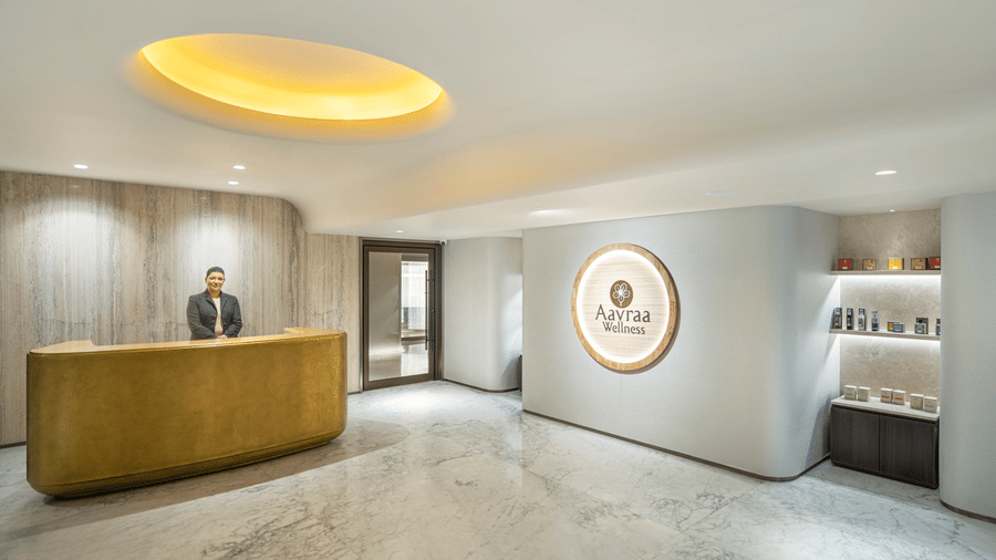 A warmly lit reception area of Aavraa Wellness at The Residency Towers Chennai with a receptionist at the desk and products kept on shelves next to the logo on the wall