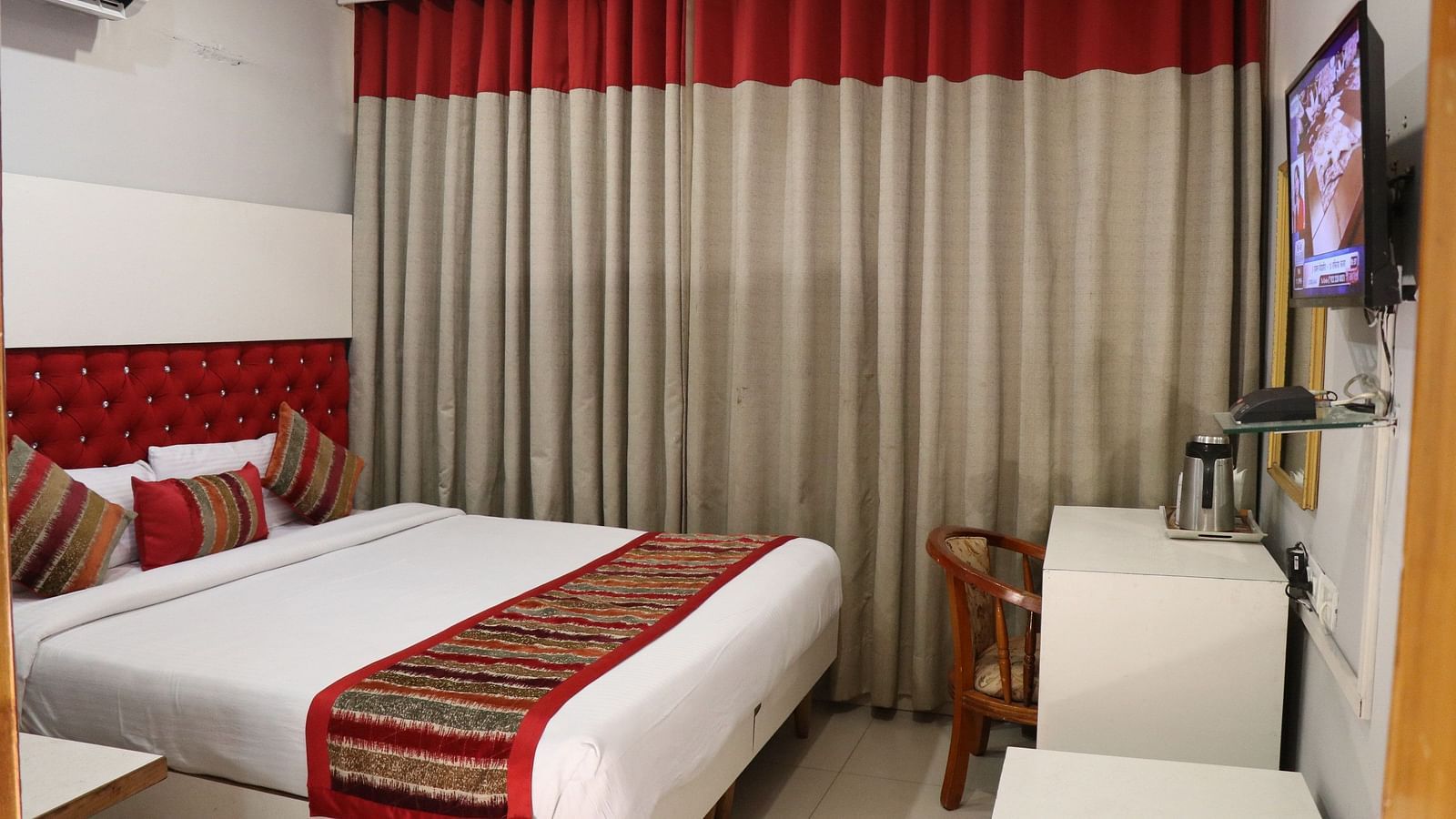 A hotel room with a plush bed, layered curtains with a red valance, a TV, a wooden chair, and an AC unit, illuminated by soft lighting. - Hotel Diamond Plaza, Chandigarh.