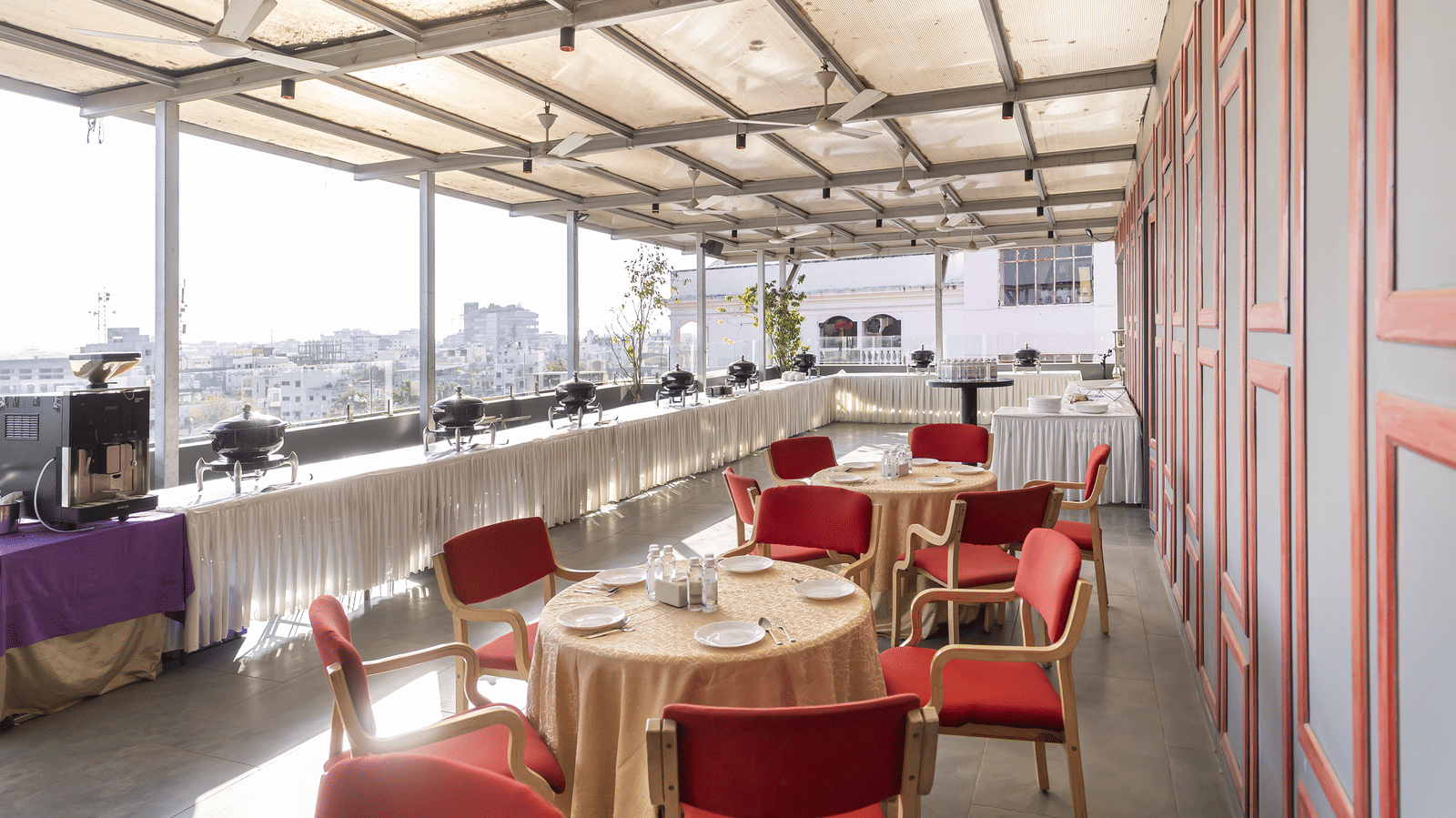 An outdoor seating area with round tables, red chairs and a roof covering with a city view at Infiniti Hotel, Indore.
