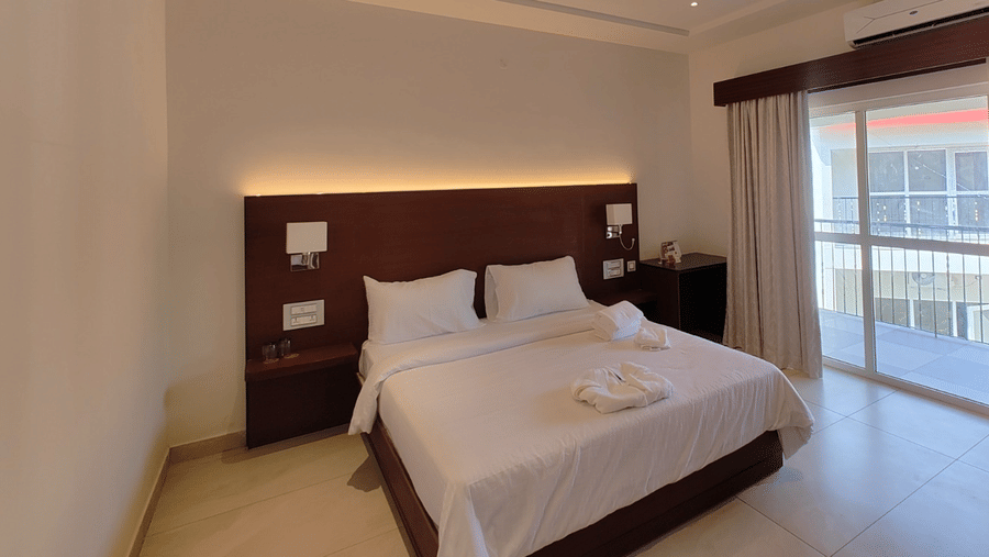 A hotel bedroom featuring a double bed with white linen, a dark wood headboard, and a sliding glass door opening onto a balcony | Lords Sahyadri Resort, Shivamogga