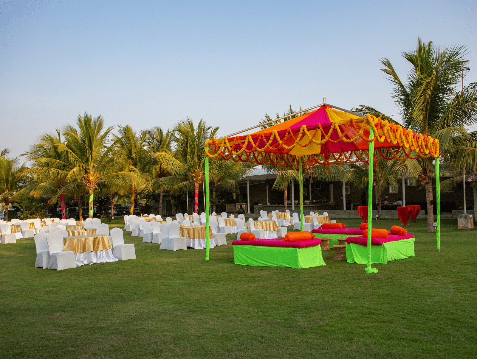 An outdoor event setup with an orange and yellow tent hosting green and red seating, and numerous white chairs on a grassy lawn - Grande Bay Resort & Spa, Mamallapuram