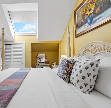 A room with double bed with designed pillows on top, wall art, and cozy yellow decor at Hartstone Inn.
