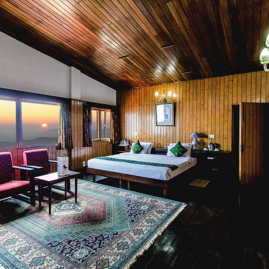 An image of a cosy room with warm wooden interiors, featuring a neatly arranged bed, sofa chairs, and a table, complemented by large glass windows offering a beautiful view of the mountains and the sun - Central Gleneagles Heritage Resort, The Mall Road, D
