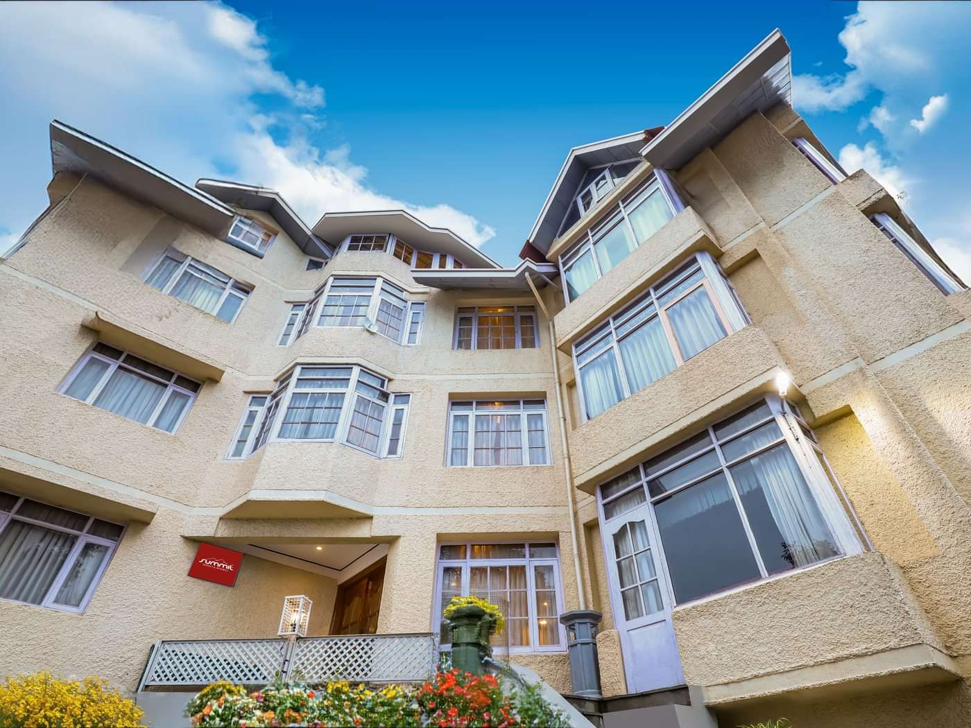 Facade view of Summit Le Royale Hotel, Shimla featuring multiple storey building. Each floor has large windows and the entrance is decorated with plants and flowers. There is also a backdrop of blue and bright sky.