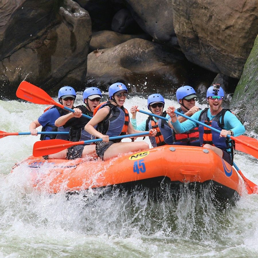 alt-text Another group of people in blue life jackets and helmets navigating through white water rapids in an orange raft, with large rocks and turbulent water around.
